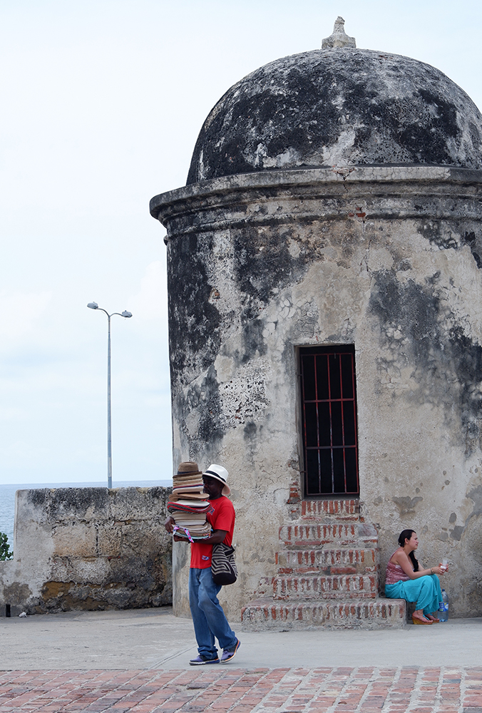 What to do and see Cartagena's Walled City and other Colombian basics: a vendor carrying a high tower of hats positioned in front of an old tower at the top of the city wall