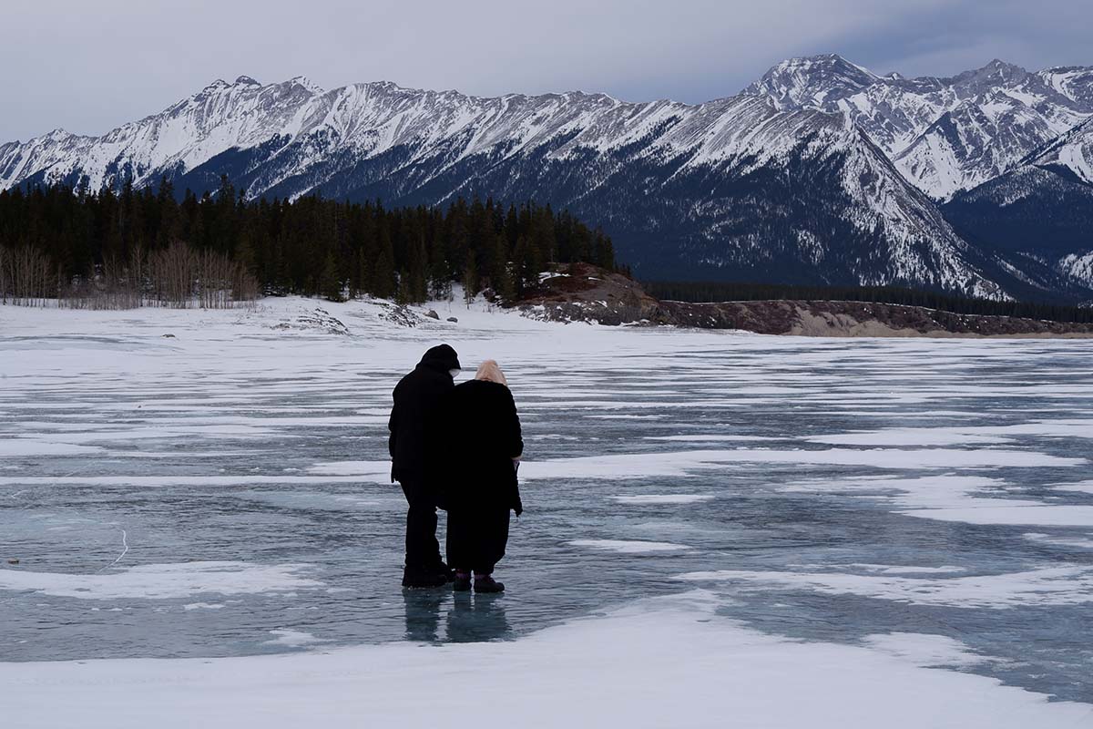 Five things to do in Banff for non-skiers: Abraham Lake wide angle view with the mountain in the back and two people in the center looking down at the ice